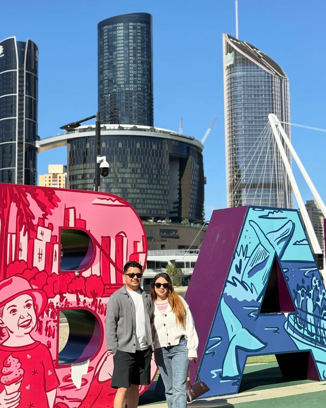 Suyog and Susmita beside the colourful Brisbane sign, with city buildings and a bridge behind them